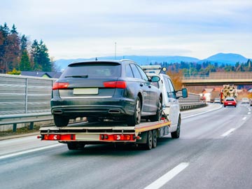 Ein Abschleppwagen von Unfallwagen Hannover transportiert ein schwarzes Auto auf einem flachen Anhänger entlang einer Autobahn, mit Bäumen und Bergen im Hintergrund.